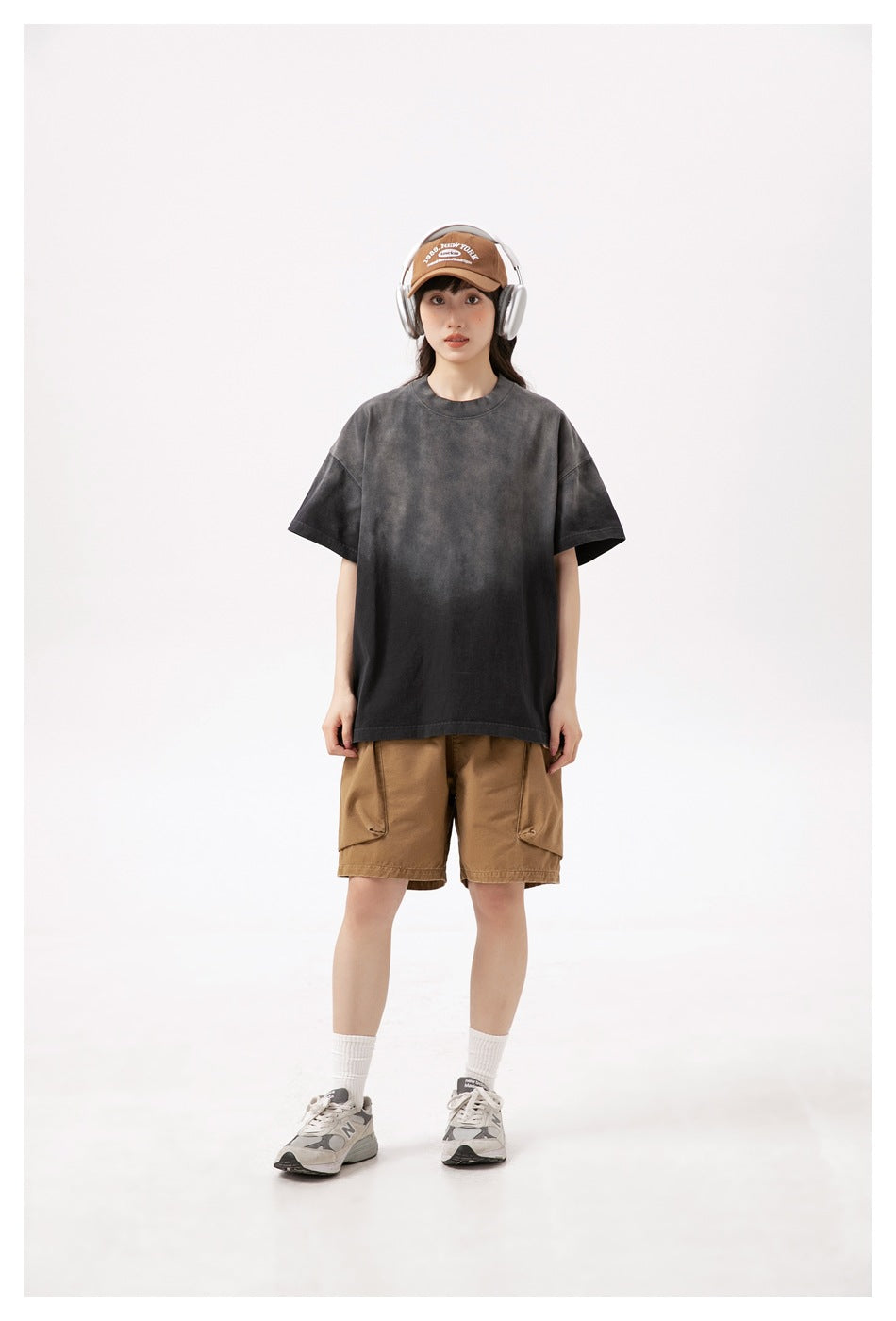 Model wearing brown cargo shorts and an oversized charcoal T-shirt, with a brown cap, headphones, white socks and sneakers.