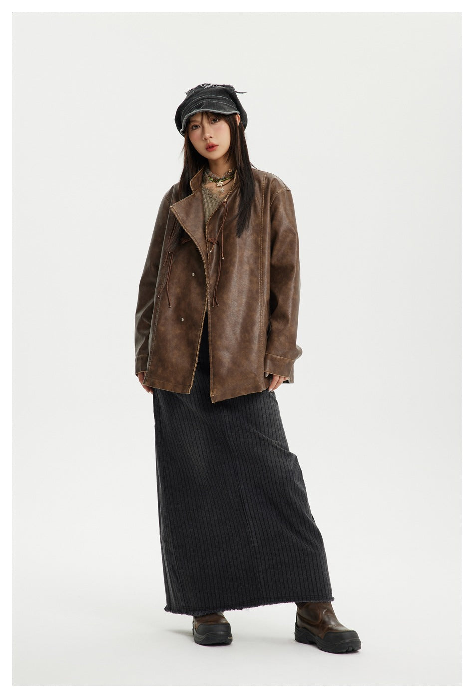 Model wearing a brown faux-leather-look jacket open over a layered top with a long gray ribbed skirt and cap