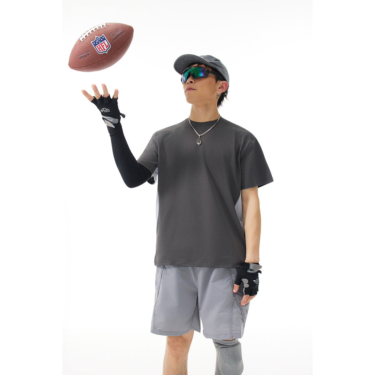 Model wearing a charcoal gray T-shirt and light gray shorts with a cap and sunglasses, tossing a football.