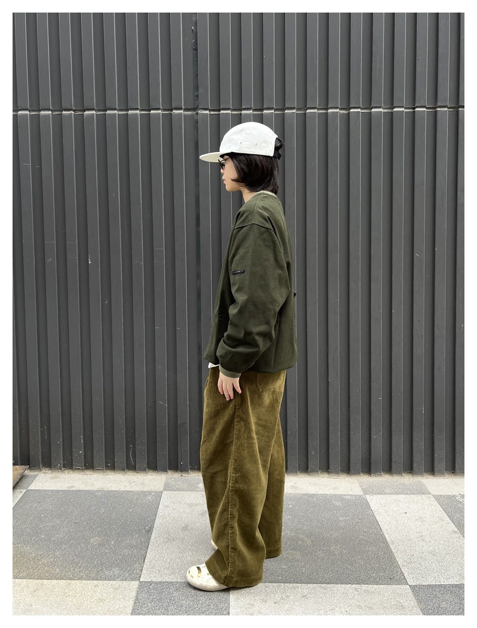 Person in profile wearing a green sweatshirt with olive corduroy pants and a white cap against a gray wall