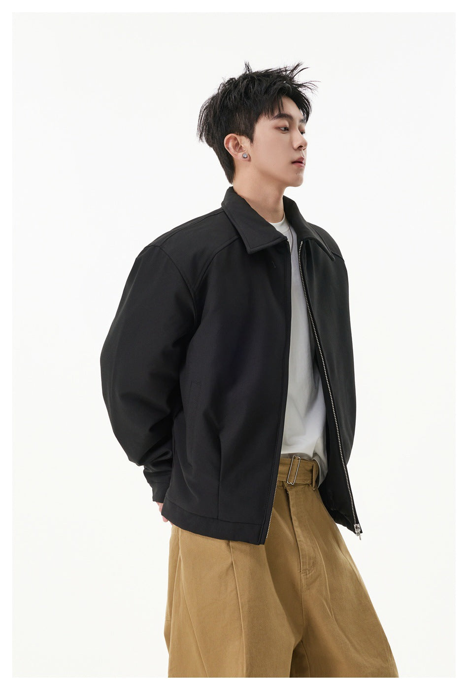 Side view of a model wearing an open black collared zip jacket over a white shirt and tan trousers.