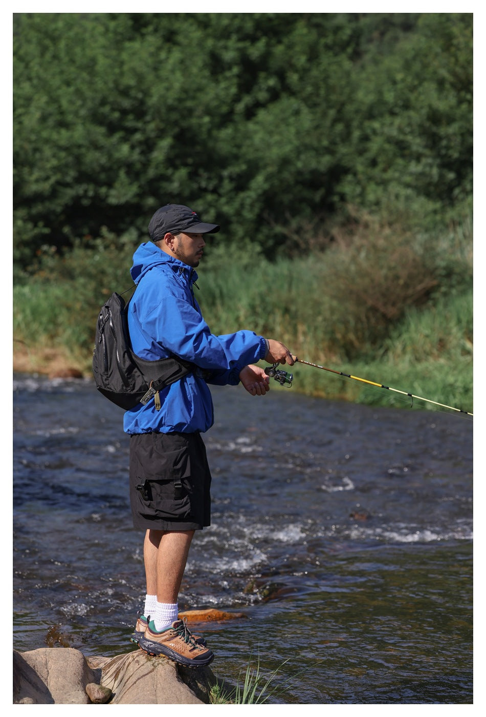 Side view of a man fishing in a blue hooded jacket and black cargo shorts beside a river.