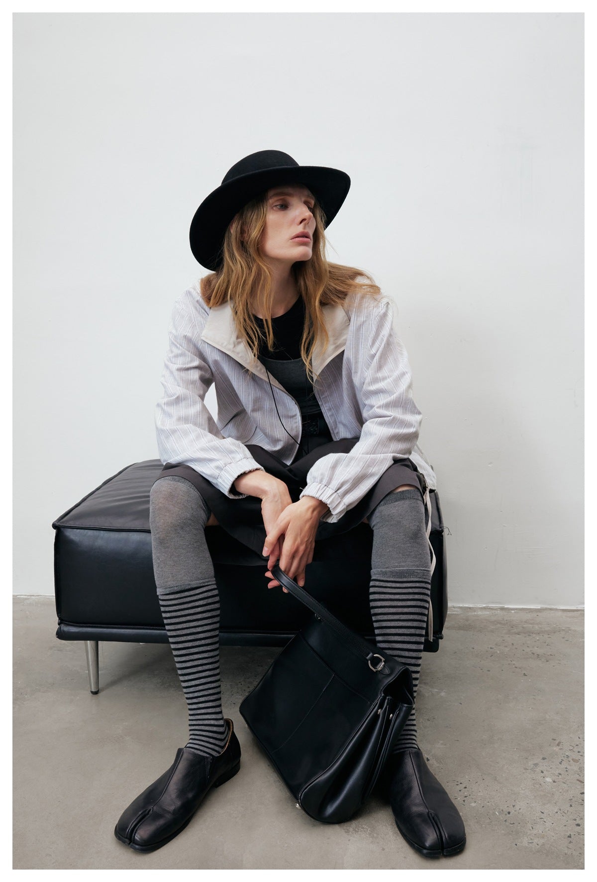 Model seated in a pale gray striped jacket with a cream collar, styled with dark clothing and a black hat.