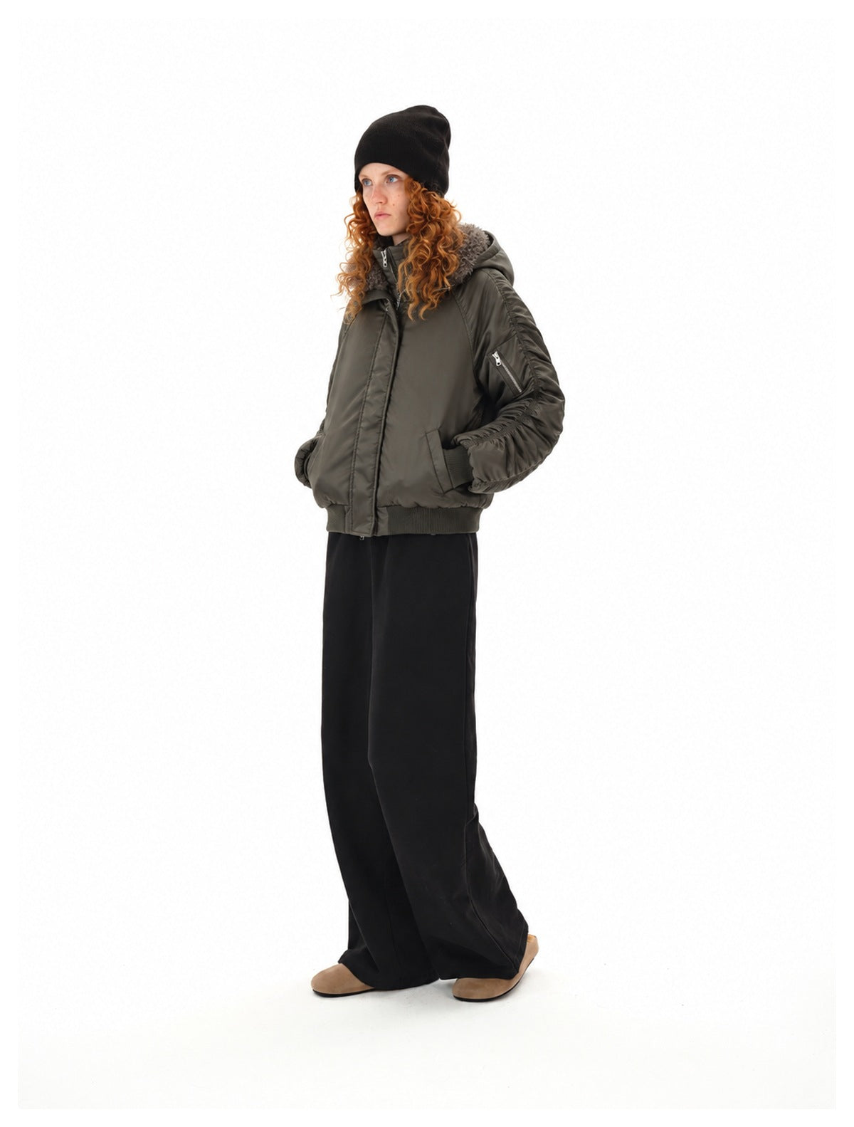 Full-length three-quarter view of a model wearing a dark olive bomber jacket with a plush gray collar and black trousers.