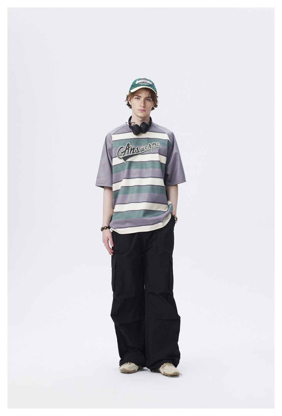 Model wearing gray striped raglan T-shirt with black cargo pants, green cap and headphones around neck, standing front view