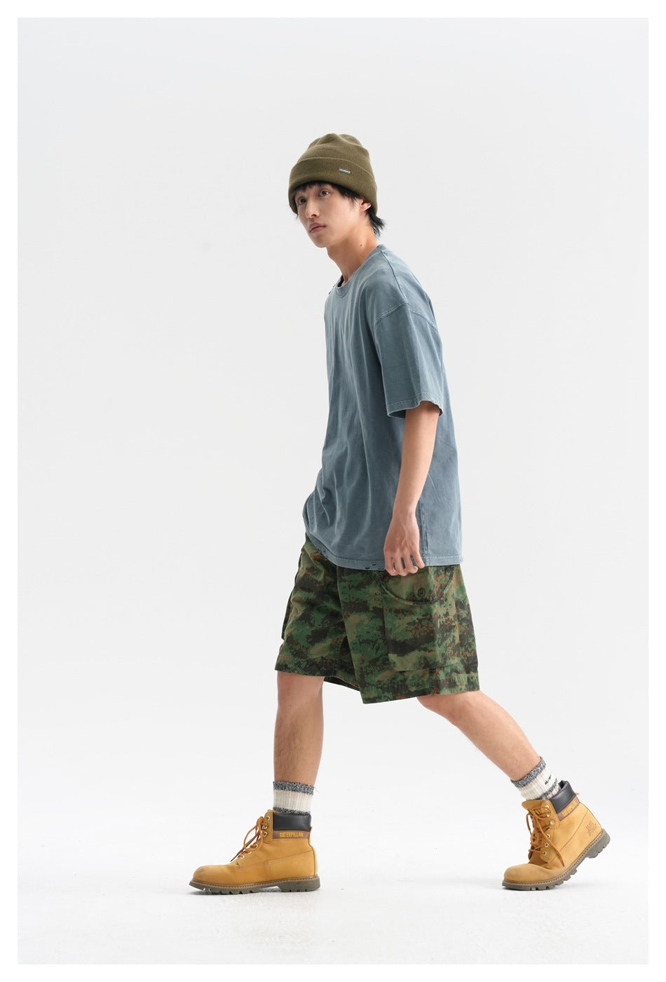 Side view of model wearing a faded blue-gray T-shirt, green camouflage cargo shorts, olive beanie, and tan boots.