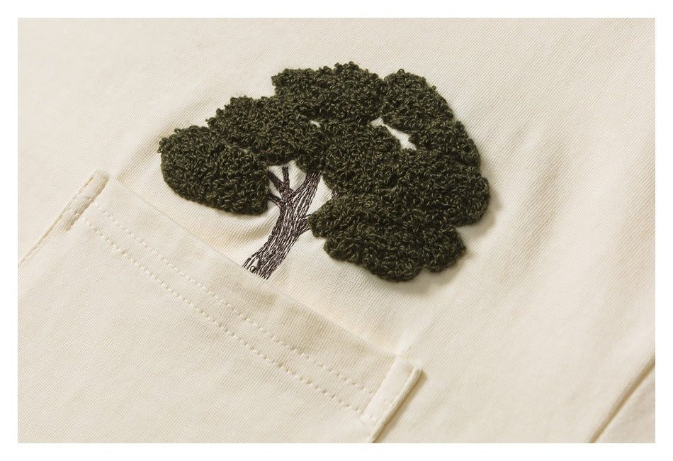 Close-up of a dark raised embroidered tree above the pocket on an ivory T-shirt.