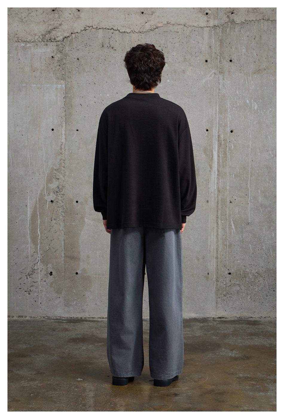 Rear view of a model wearing an oversized plain black crewneck and loose gray trousers.
