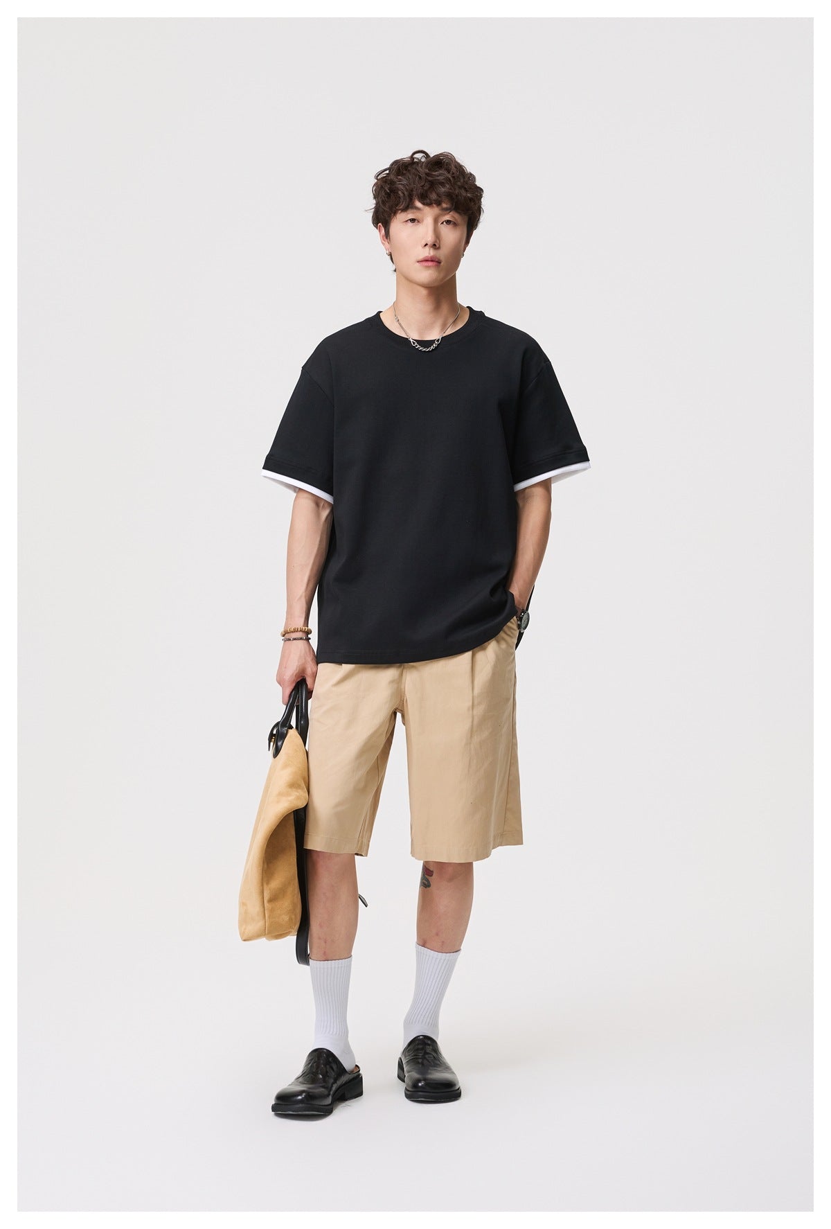 Model wearing black layered-sleeve crewneck T-shirt with khaki shorts, holding a tan bag
