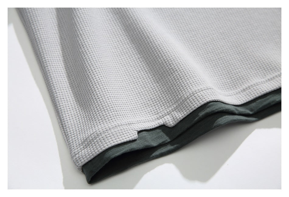 Close-up of the stitched hem of a light gray waffle-knit shirt, with a dark green layer visible underneath.