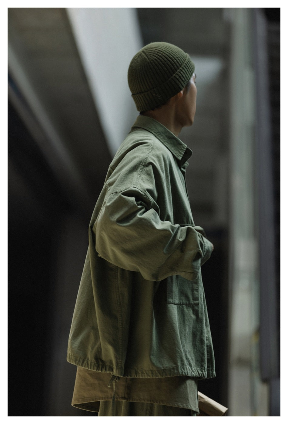 Rear-side view of a model wearing an olive-green oversized jacket and dark green ribbed beanie.