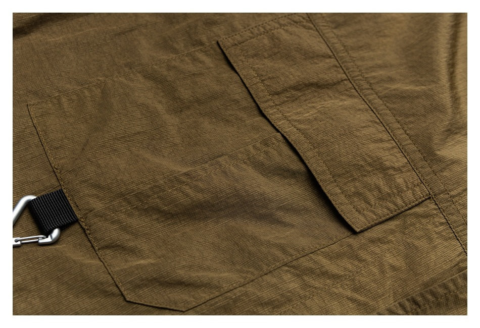 Close-up of olive-brown garment fabric with stitched panels, a flap pocket, and a small metal clip.