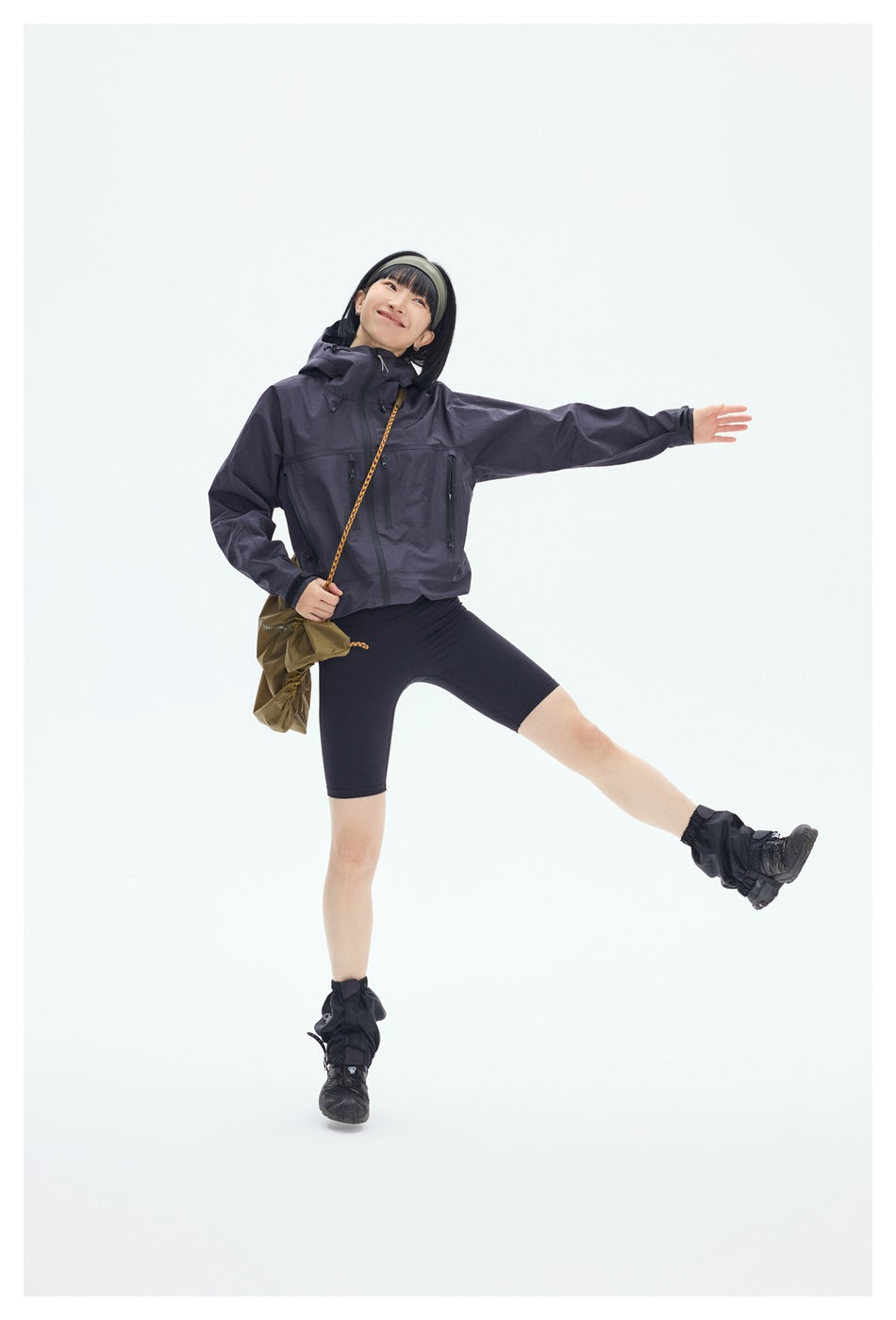 Model poses in a dark plum hooded jacket with zip pockets and black cycling shorts, one leg extended.
