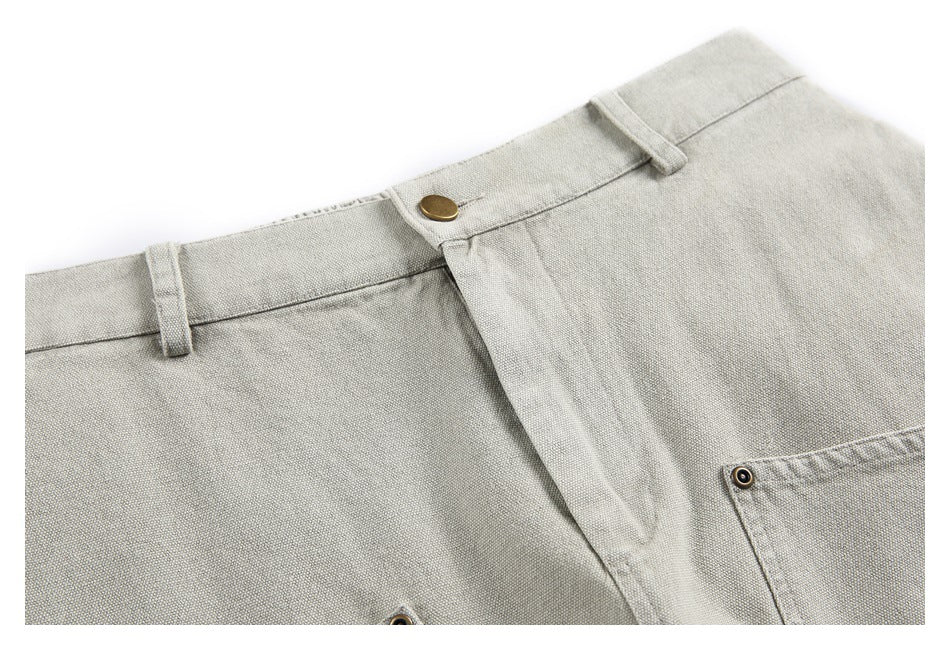 Close-up of light gray-beige bottoms showing the waistband, belt loops, brass-tone button, and front fly.