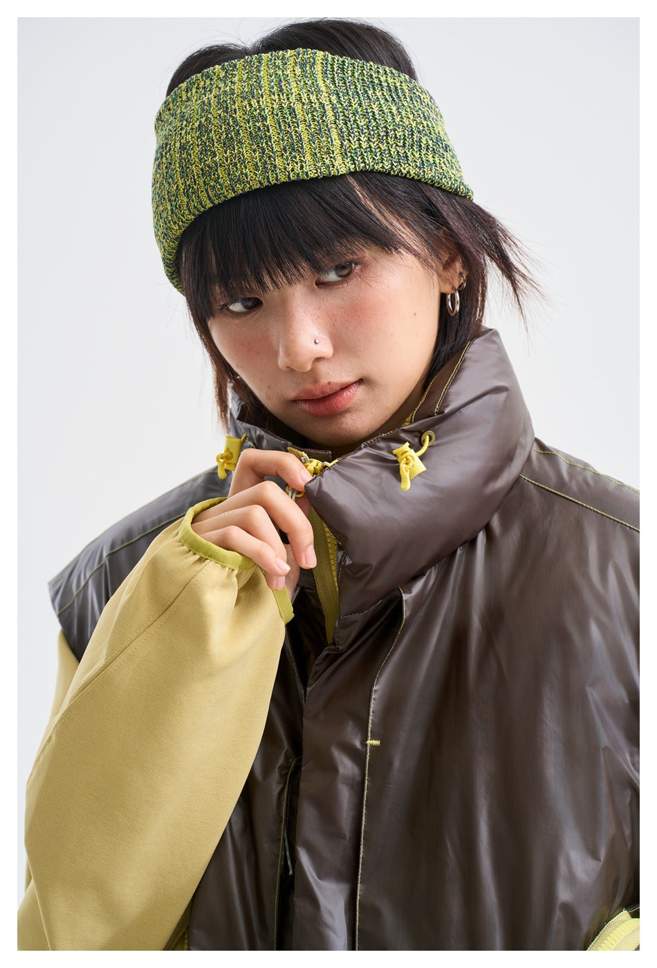 Model wearing a dark brown sleeveless vest with a tall collar and yellow fasteners over a pale yellow top.
