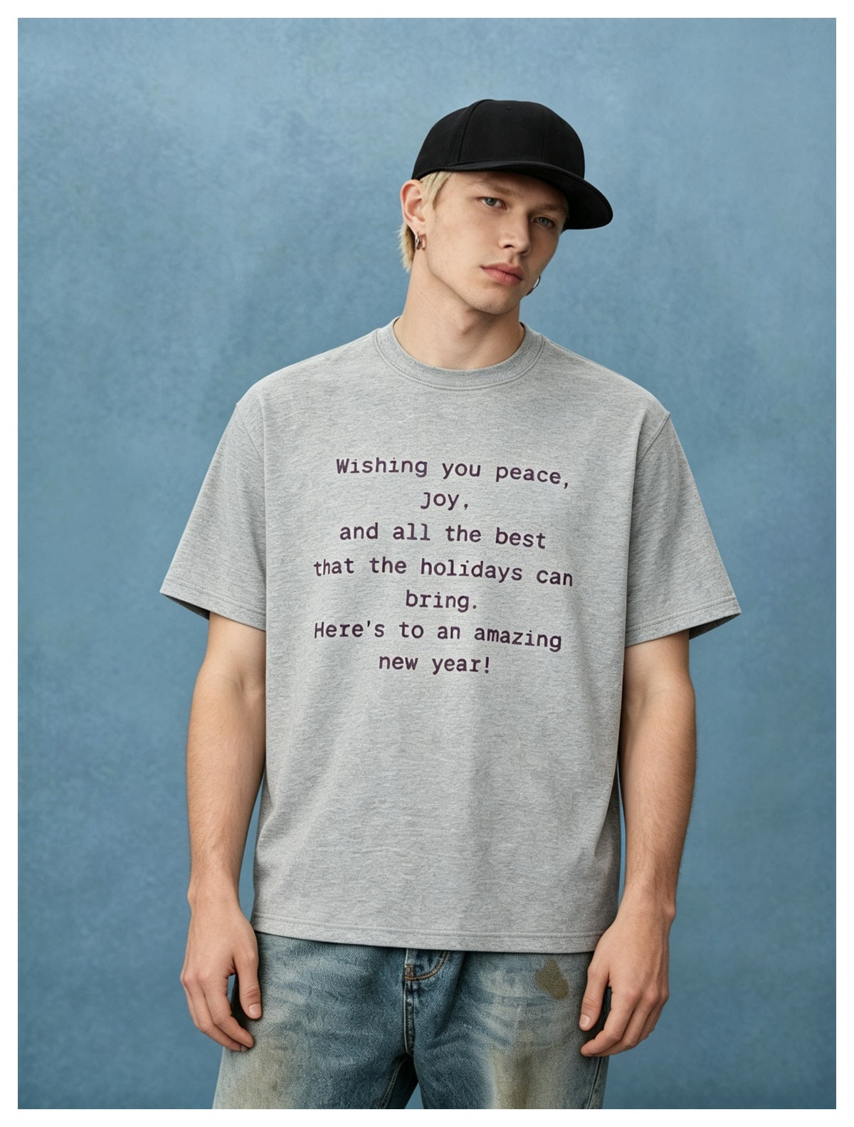 Man in a heather gray text-print T-shirt, black cap and light-wash jeans against a blue backdrop