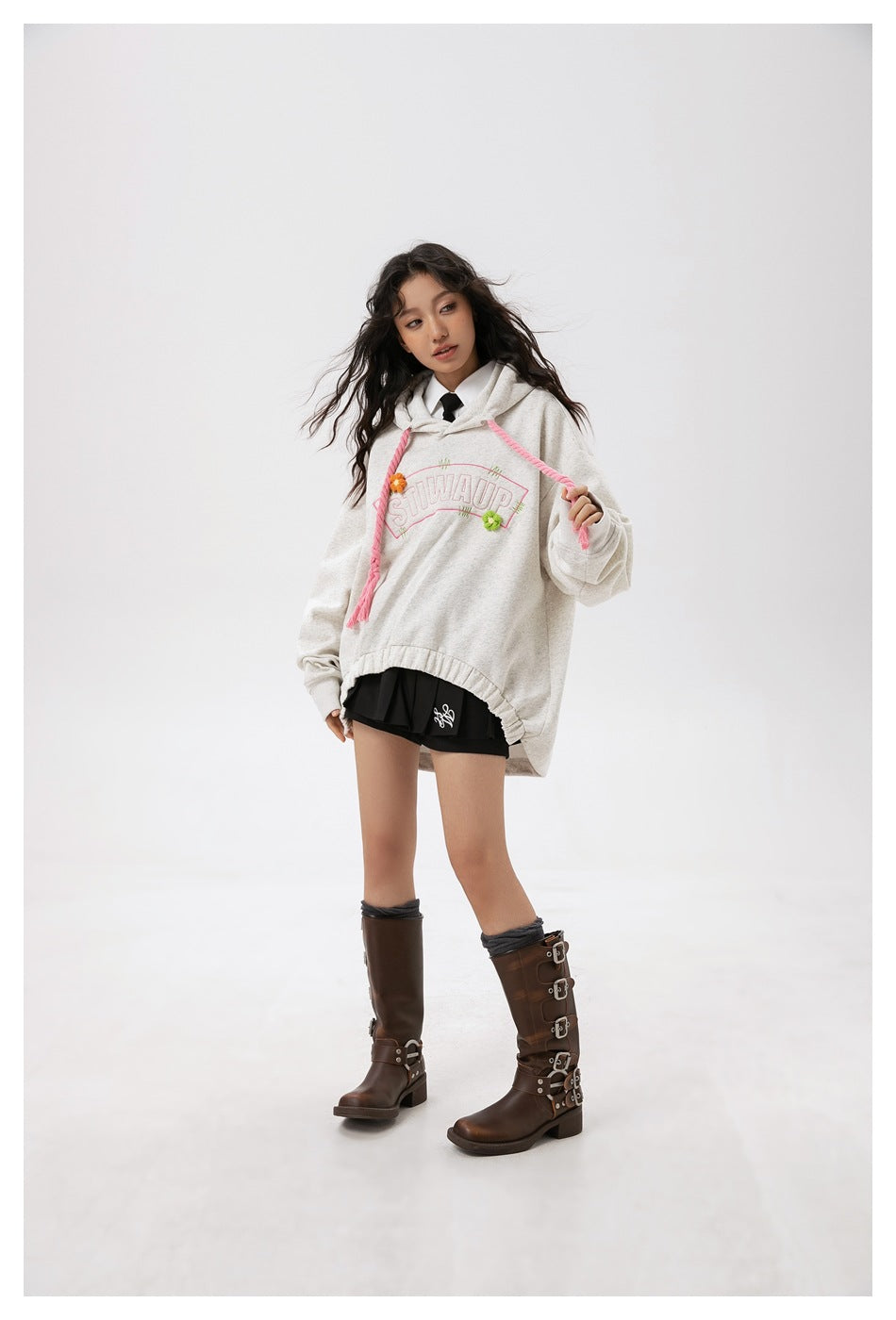 Full-length model wearing the front of an oversized light gray hoodie with pink drawstrings and floral lettering.