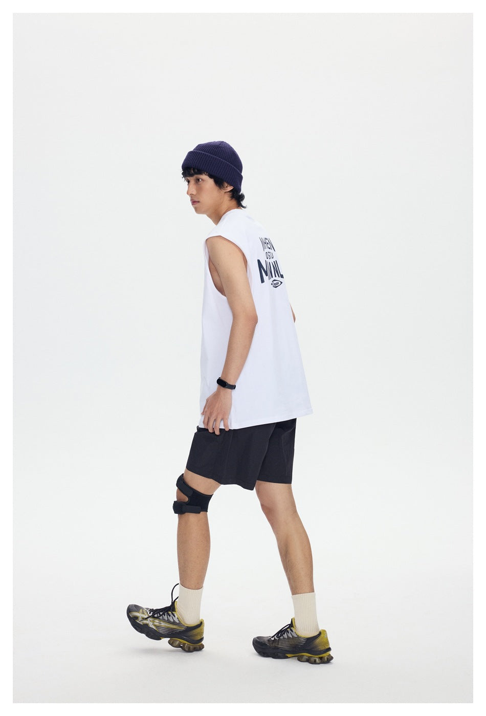 Model in side view wearing a white sleeveless top with navy back print, black shorts, navy beanie and sneakers