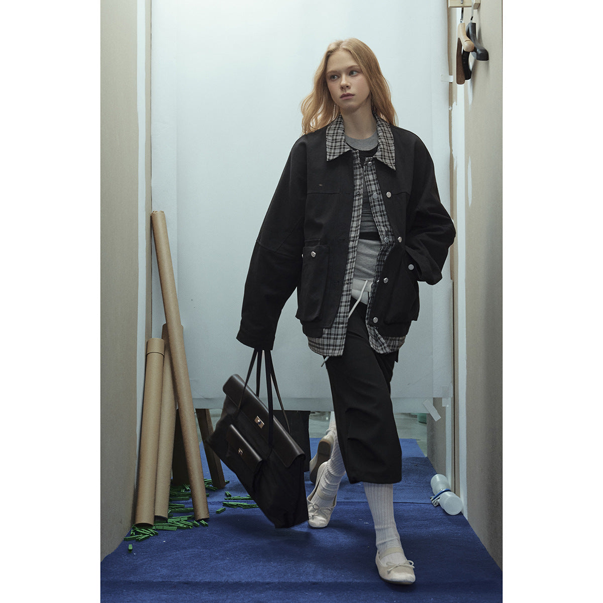 Model wearing an oversized black jacket over a long black-and-gray plaid shirt, gray top, and dark trousers.