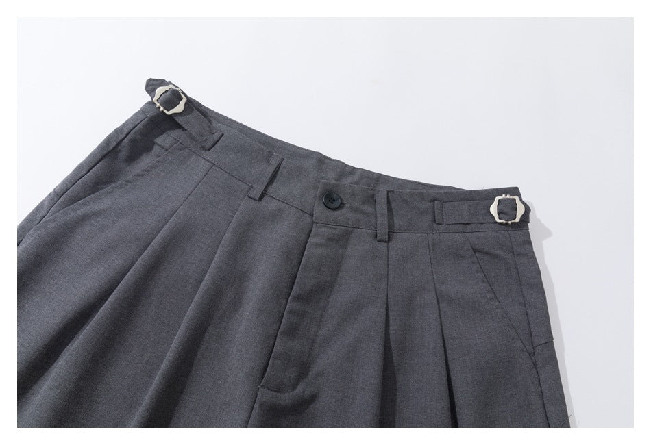 Close view of charcoal gray pleated trousers with belt loops, a black button, and adjustable buckle tabs at the waist.