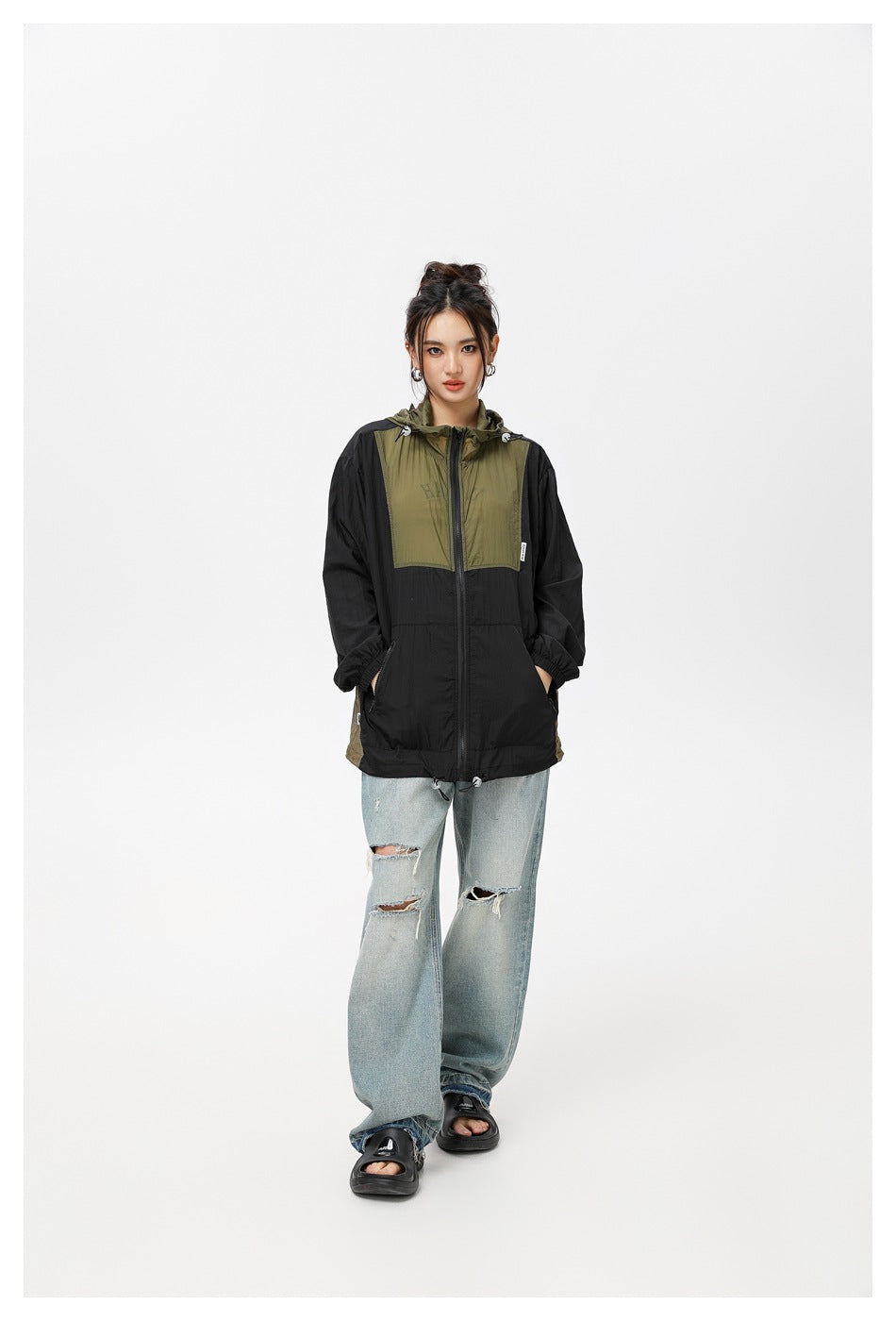 Front view of a model wearing a black and olive hooded windbreaker with loose distressed jeans.