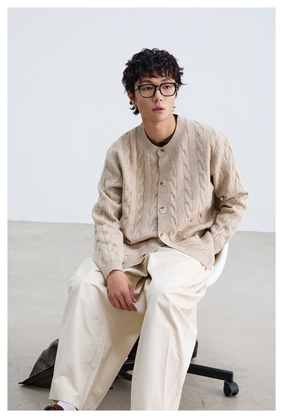 Seated model wearing a light beige cable-knit button cardigan and loose white trousers.
