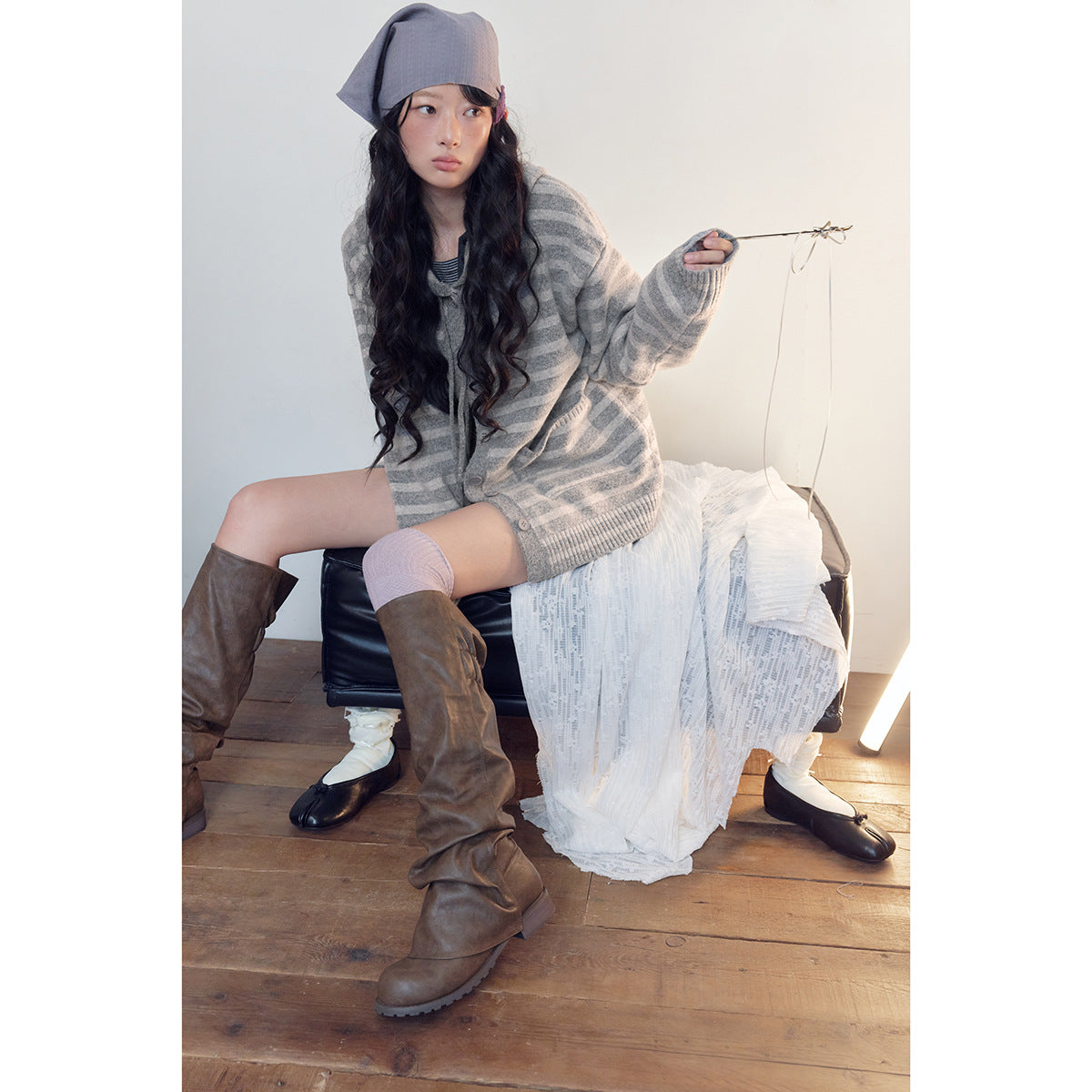 Model seated in a gray striped button-front cardigan with a hood, styled with a lavender headscarf and brown boots.