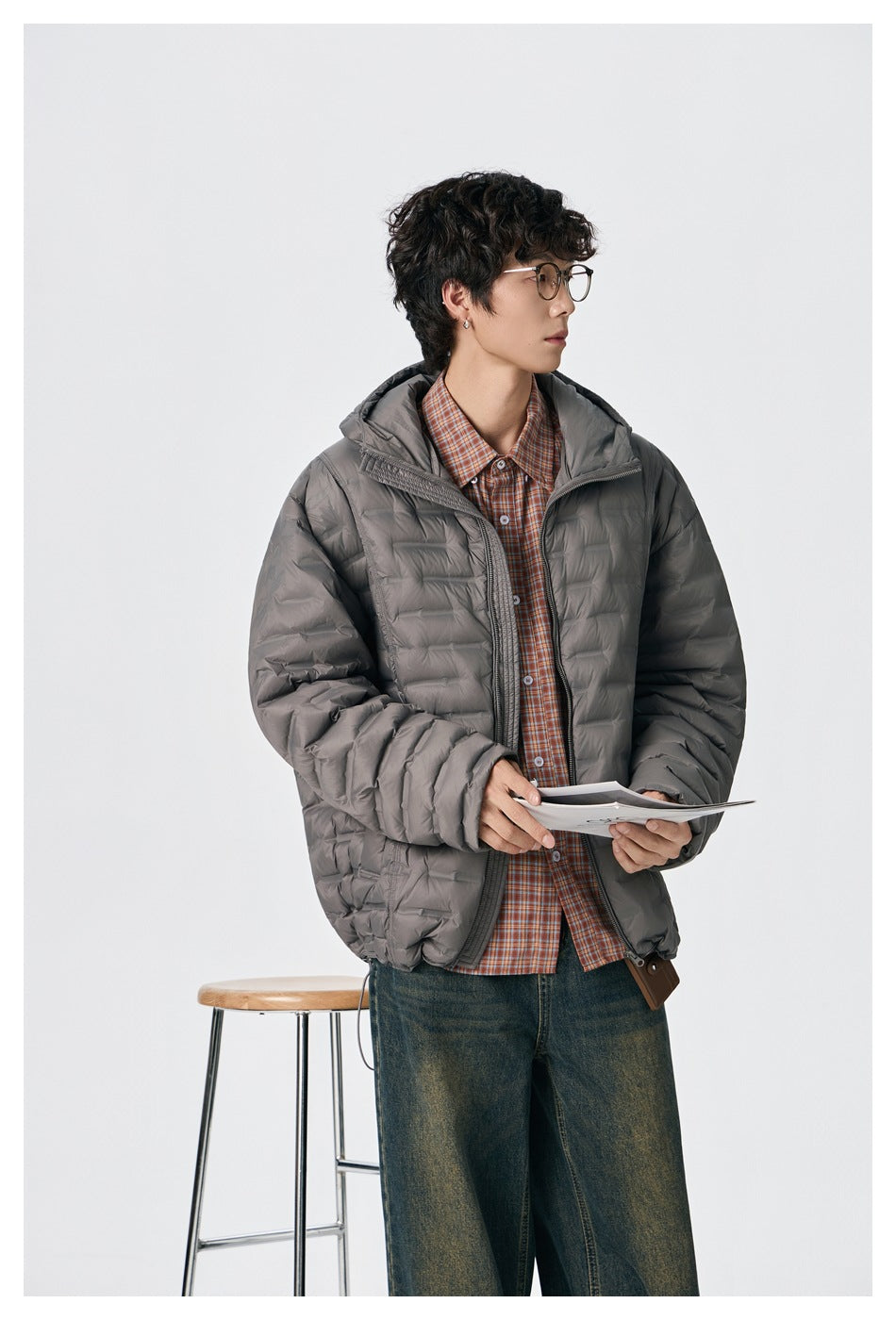 Model wearing an open gray quilted hooded jacket over a plaid shirt, holding papers.