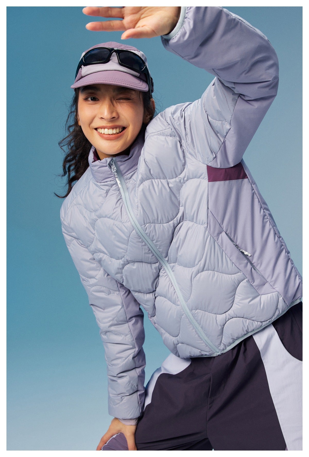 Smiling model in a pale lavender wave-quilted jacket, leaning toward the camera with one arm raised.