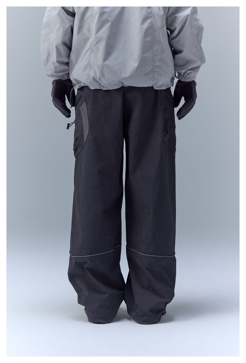 Back view of loose black pants with curved piping and cargo pockets, worn with a light gray jacket.