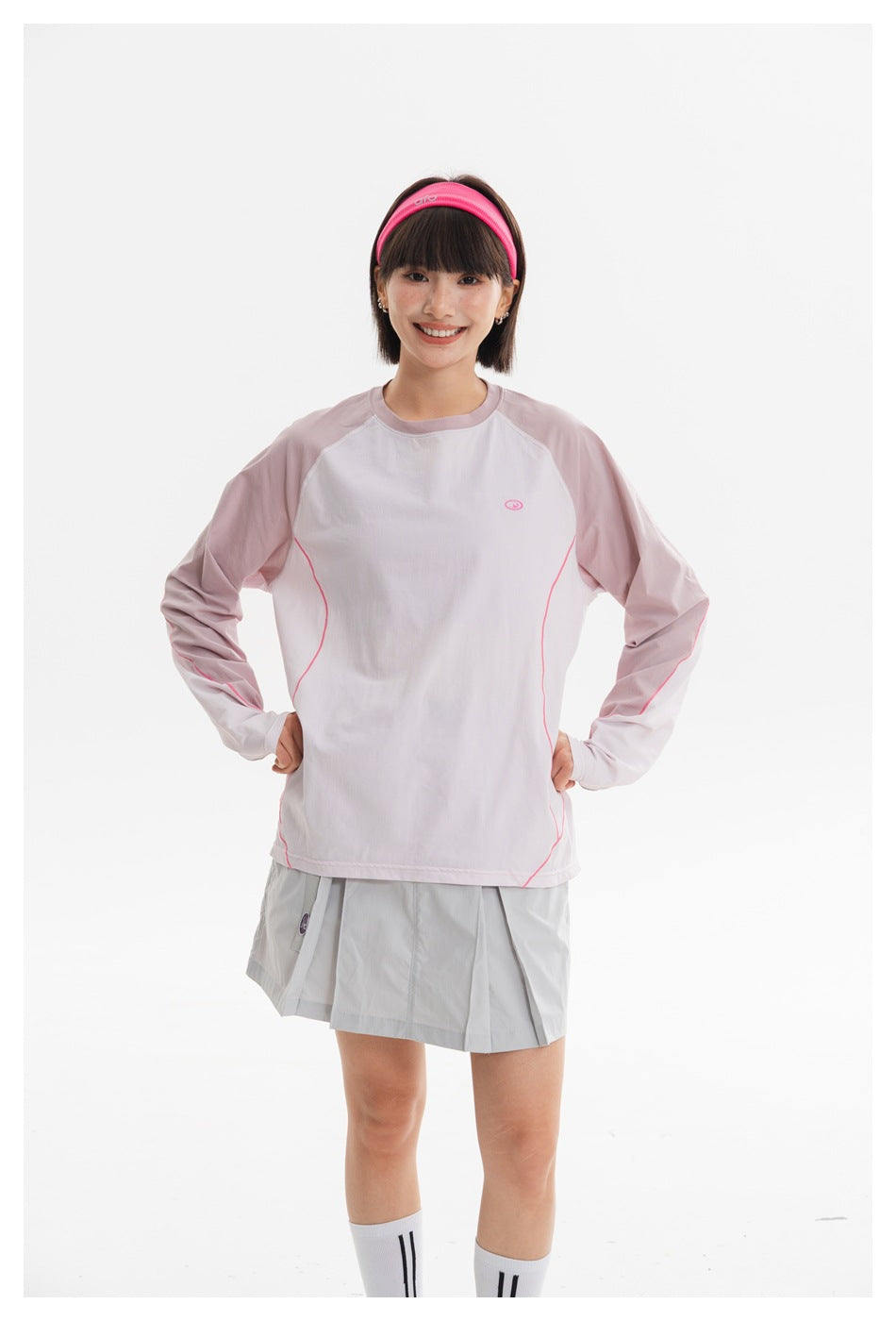 Front model view of a pale pink and white long-sleeve top with curved pink piping and a light gray pleated mini skirt.