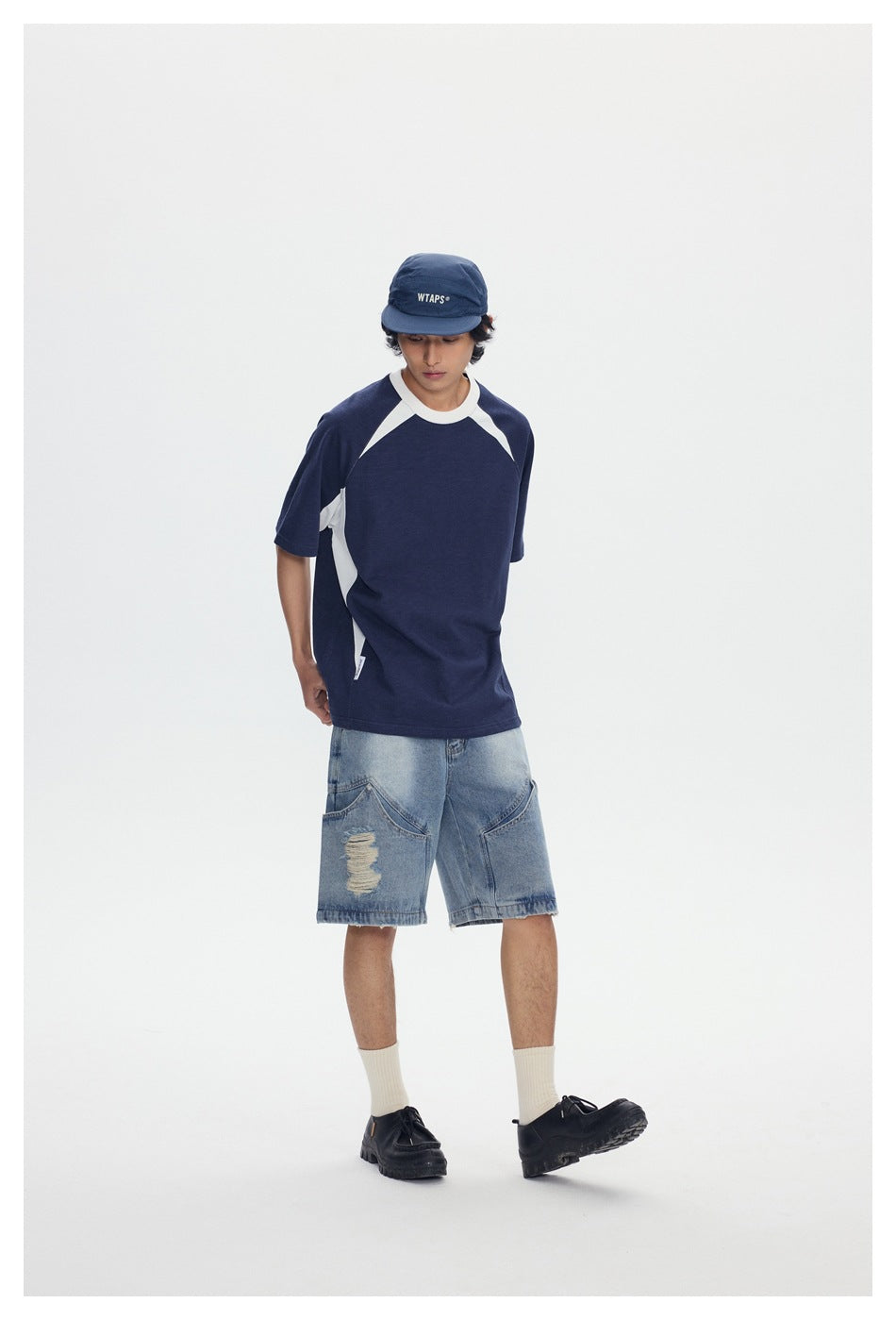 Model looking down wearing navy raglan T-shirt with white side panels, denim shorts and black shoes, angled view