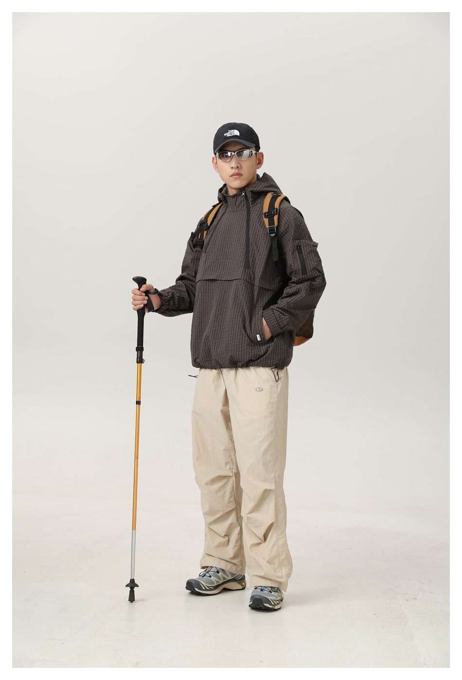 Full-length model wearing a dark brown checked hooded jacket with beige trousers and carrying a backpack and trekking pole.
