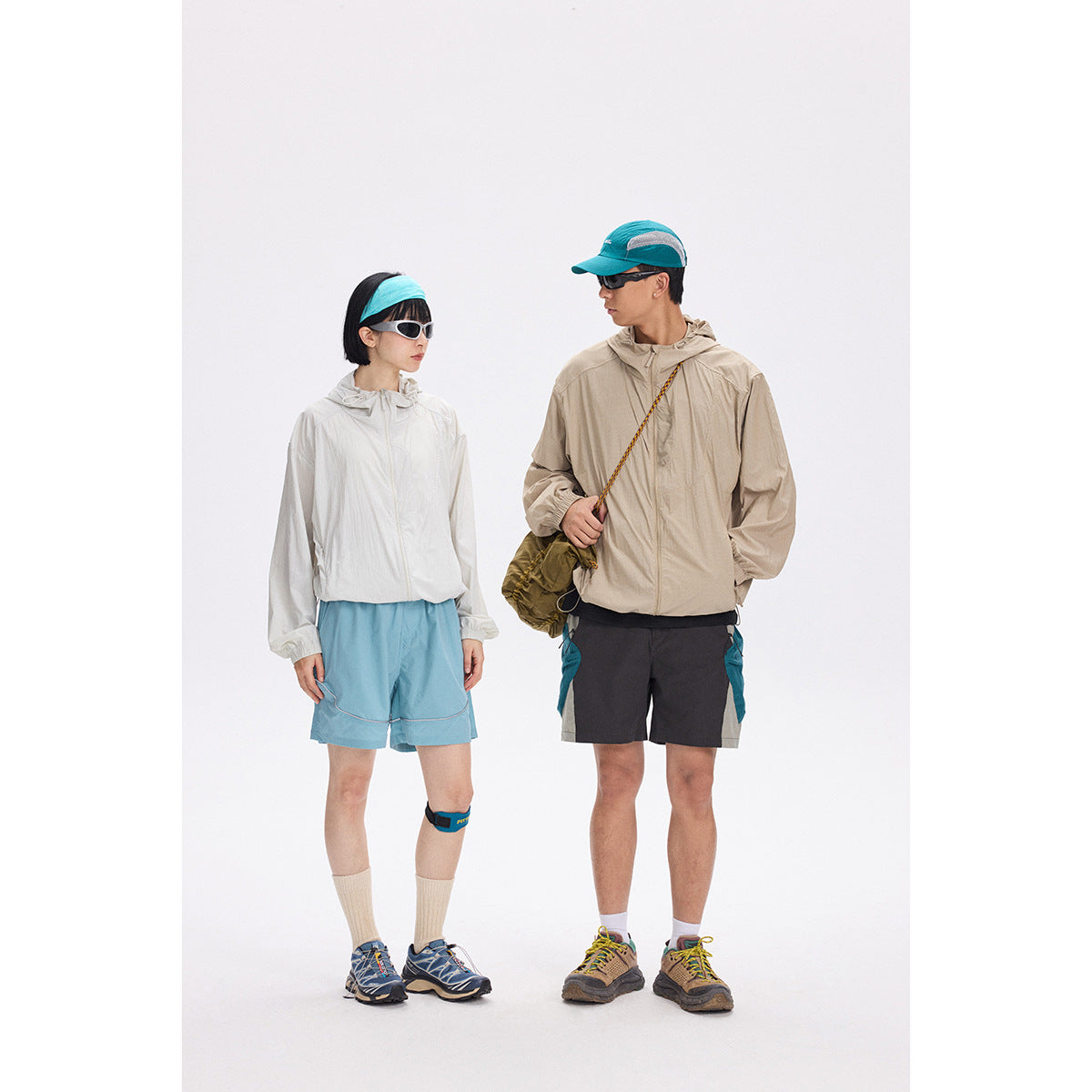 Two models wear white and beige hooded jackets with hiking shorts, caps, and sunglasses.