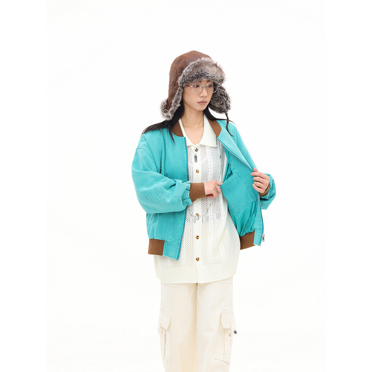 Model wearing a turquoise bomber jacket with brown trim, shown open over a cream knit cardigan and cargo pants.