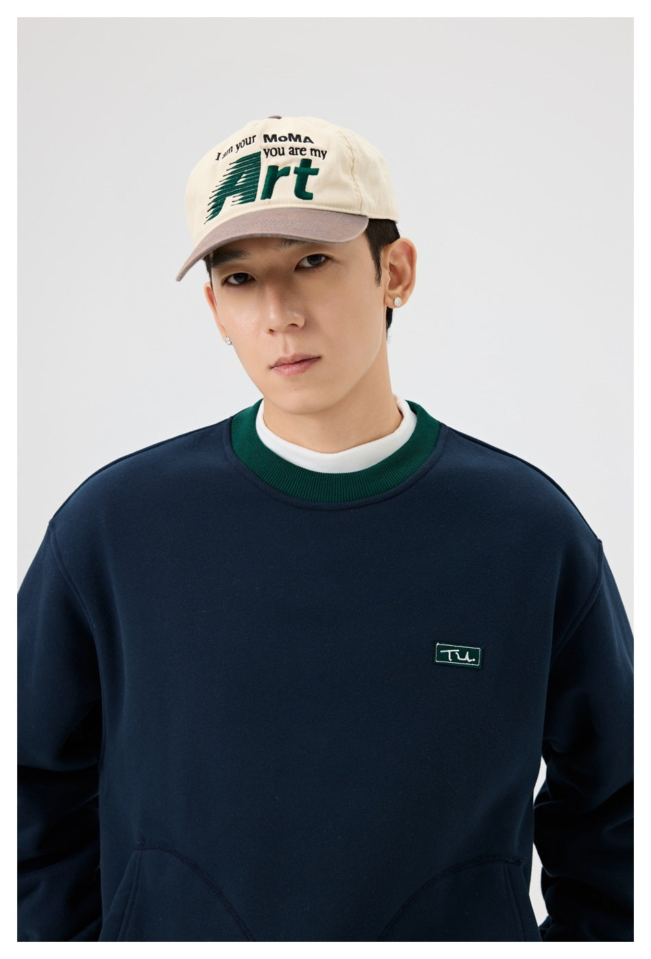 Close-up of a model wearing a navy sweatshirt with green ribbed collar and small green chest patch, with a white layer