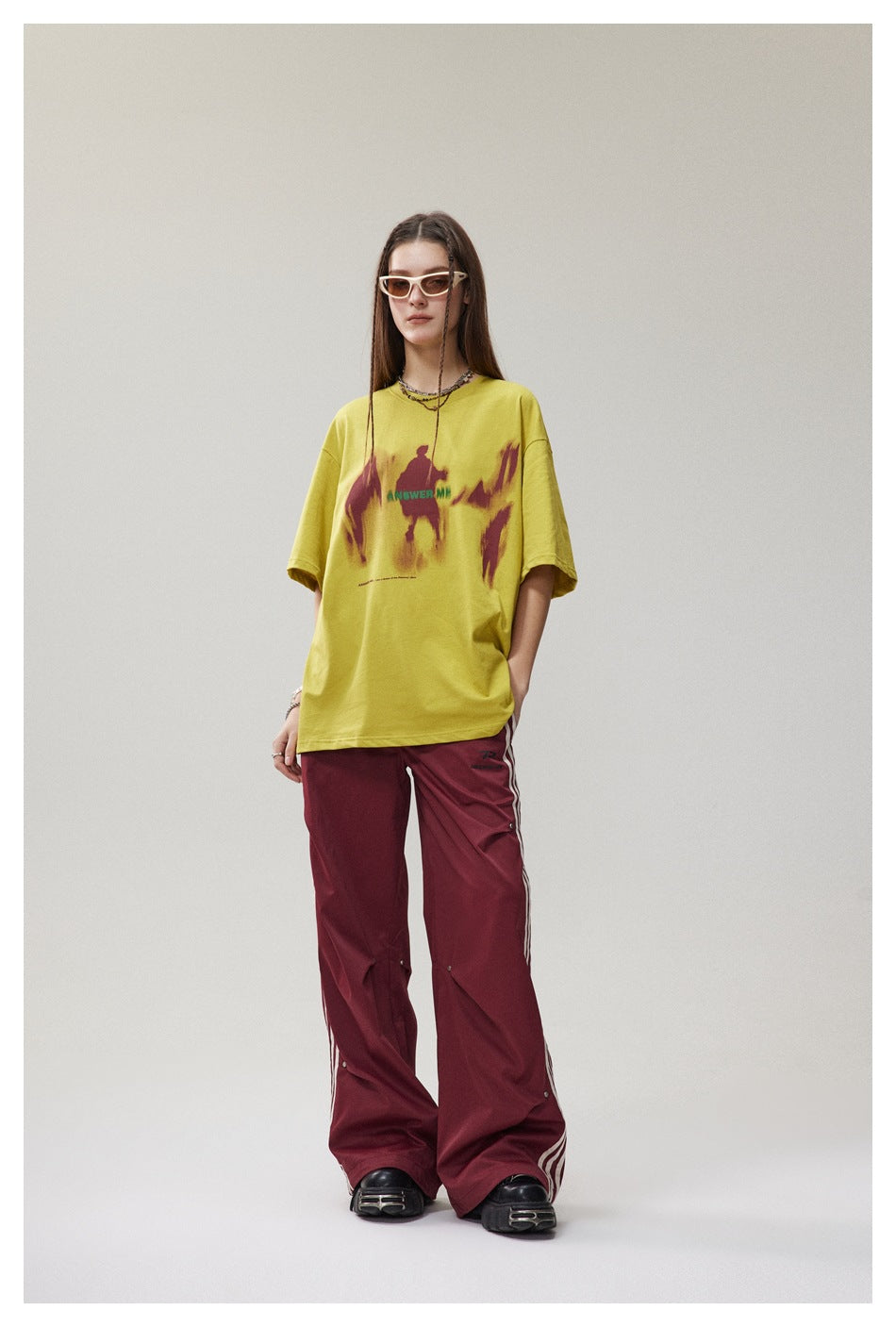 Full-length model wearing a loose yellow graphic T-shirt with burgundy abstract print and burgundy wide-leg pants.