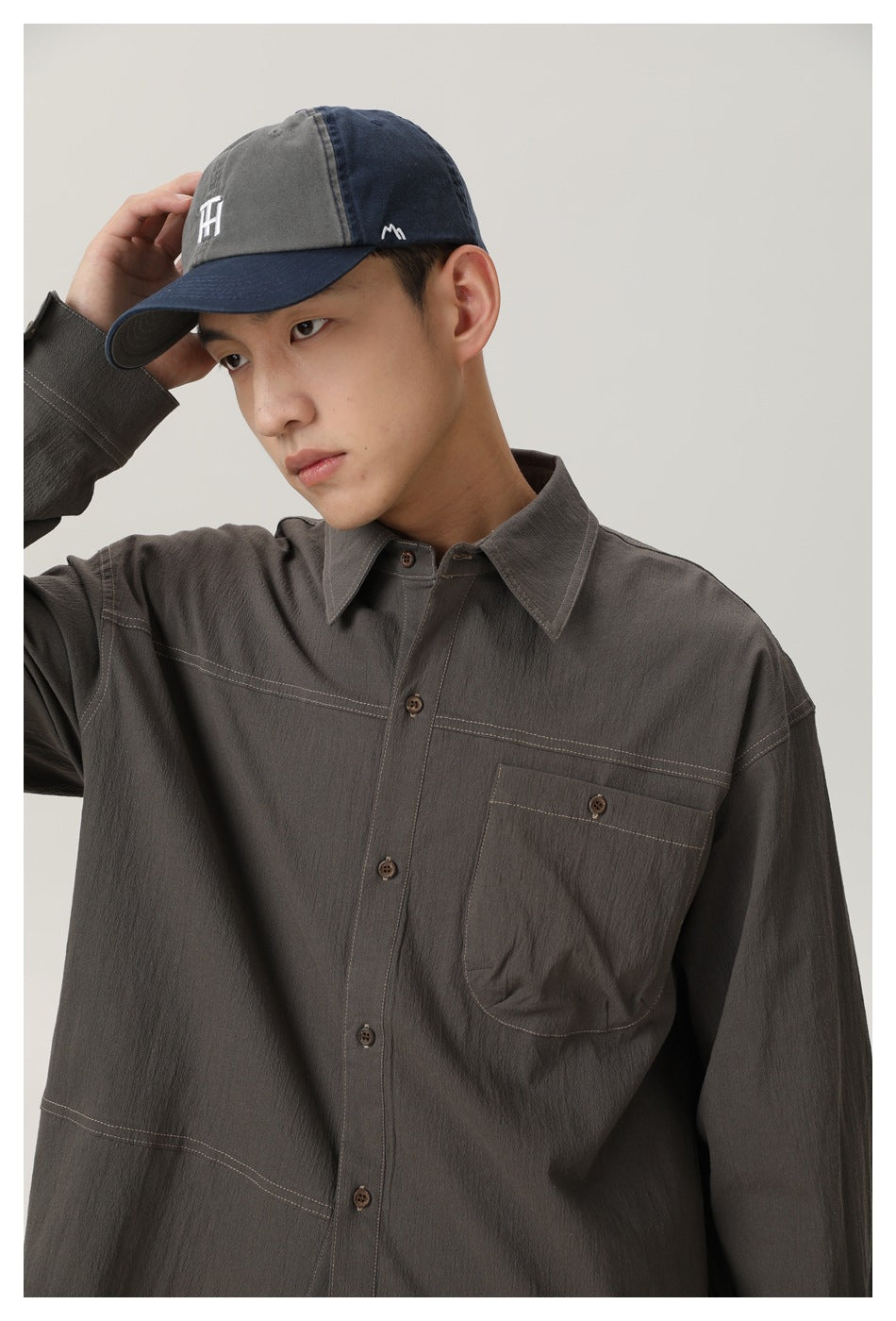 Model wearing a charcoal button-up shirt with contrast stitching and a chest pocket, plus a two-tone gray and navy cap.