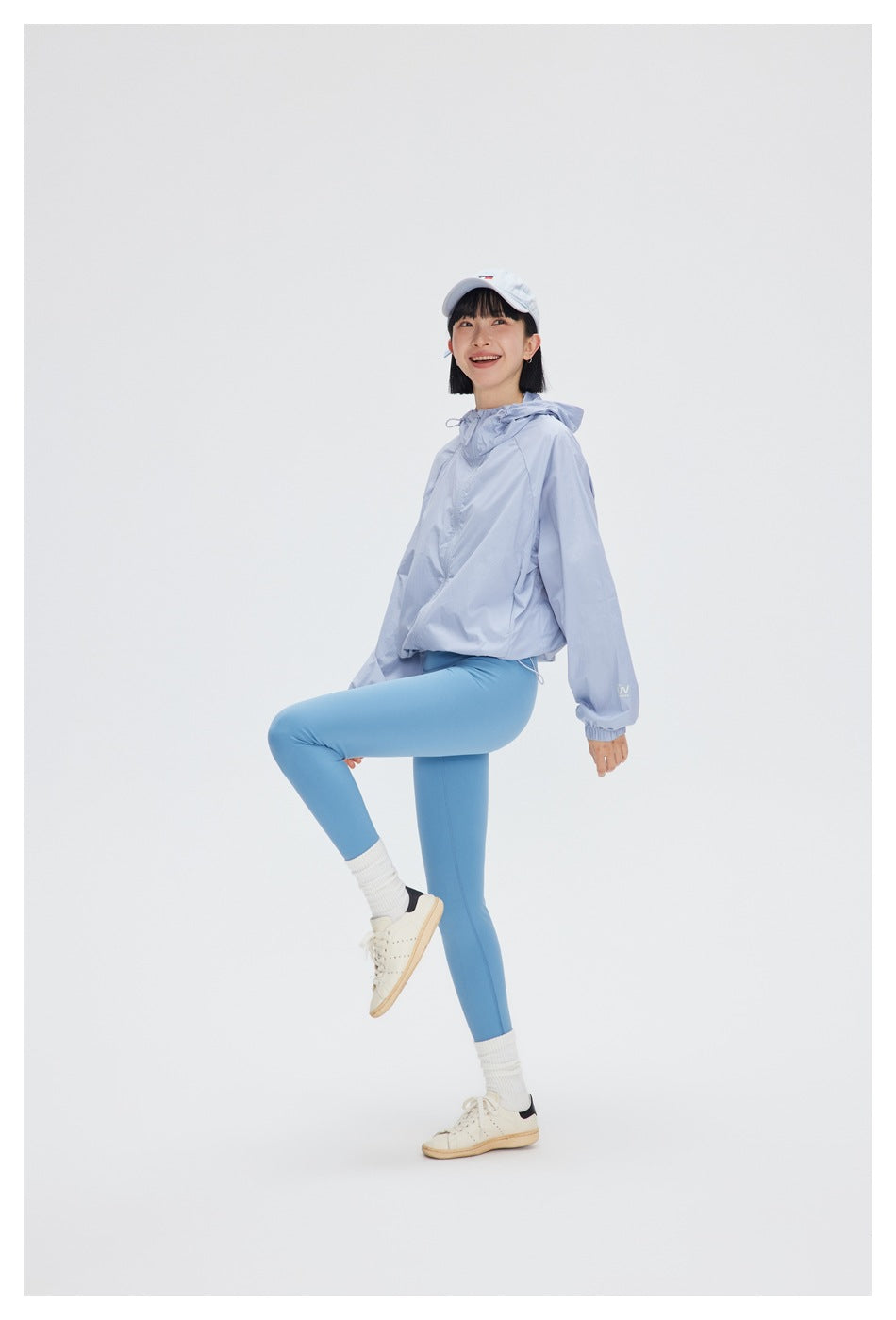 A woman poses in a pale blue hooded jacket and blue leggings, lifting one knee.