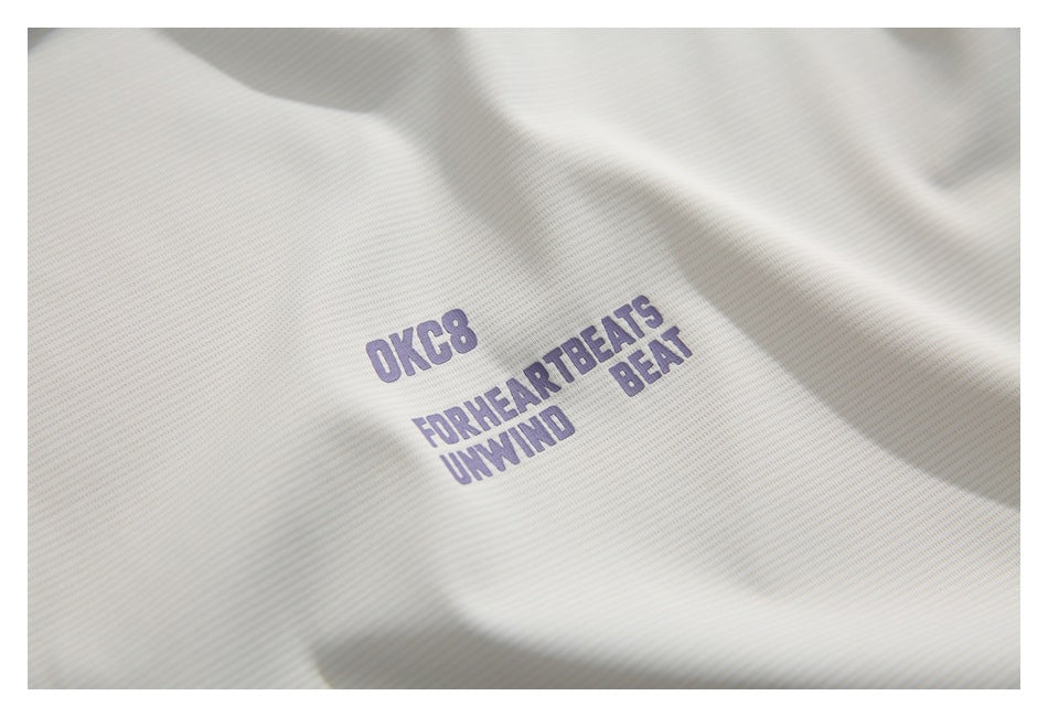 Close-up of a white textured shirt with purple “OKC8” and “FOR HEARTBEATS UNWIND BEAT” lettering.