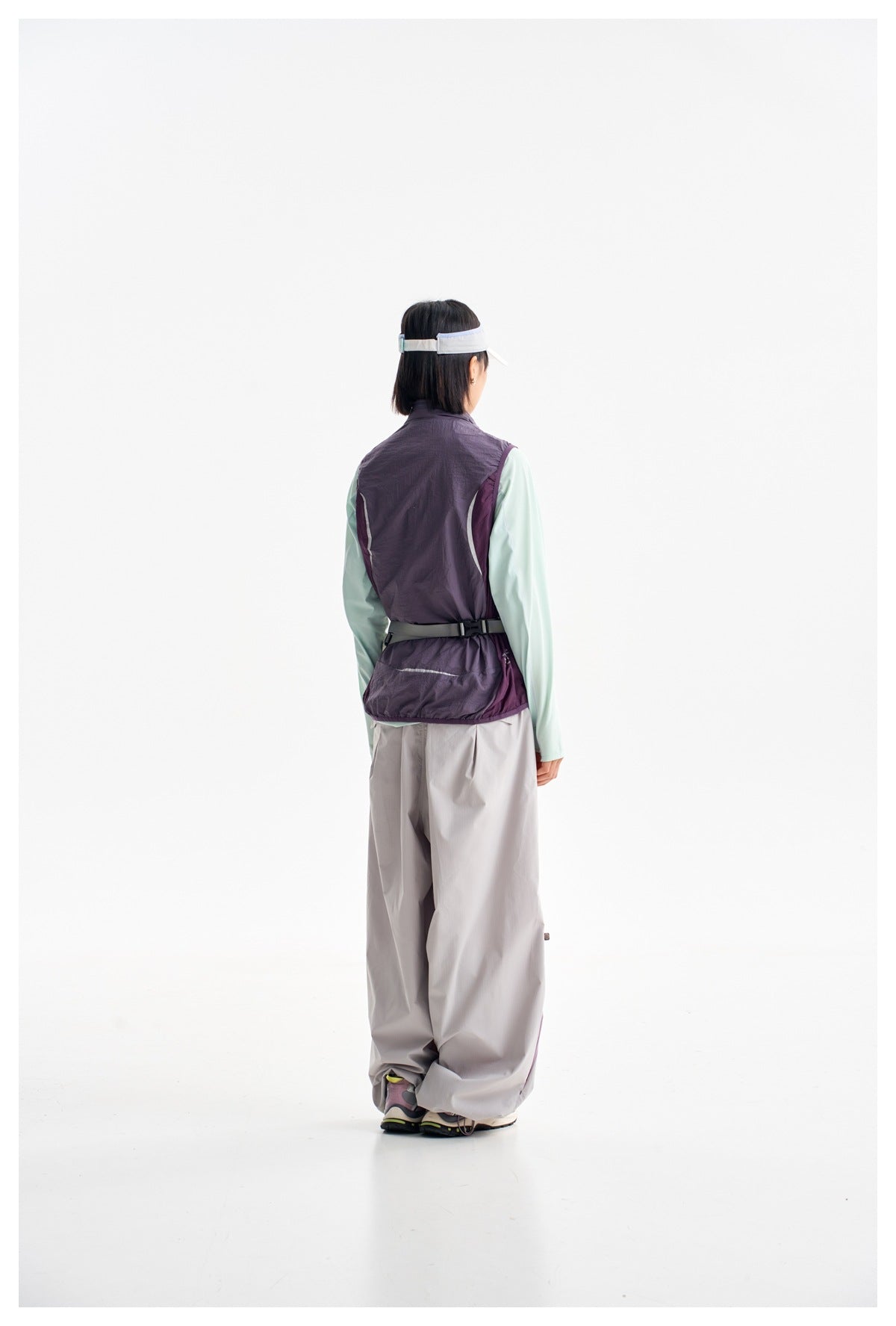 Full-length rear view of a woman wearing a plum utility vest over a mint top, loose gray pants and a white visor.