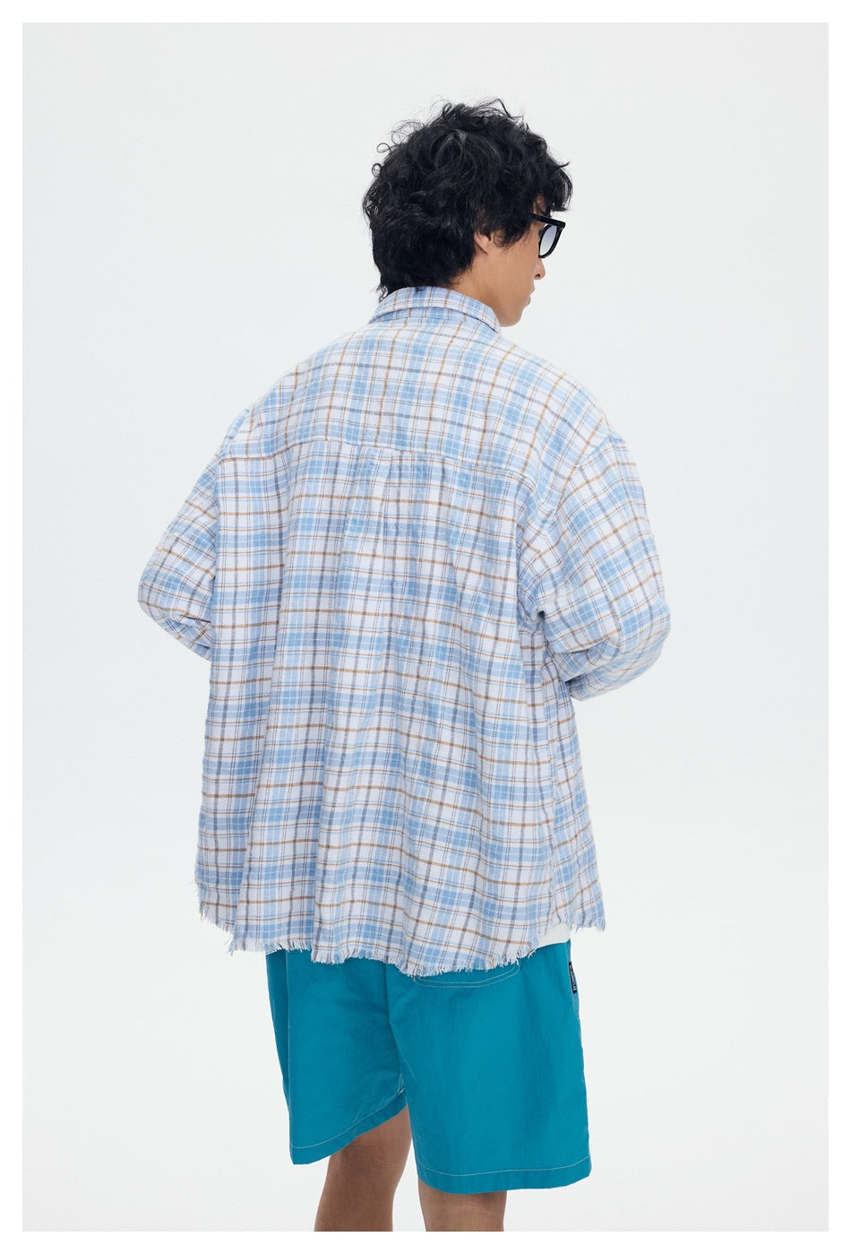 Back view of a model wearing a loose light blue and tan plaid shirt with a frayed hem and turquoise shorts.
