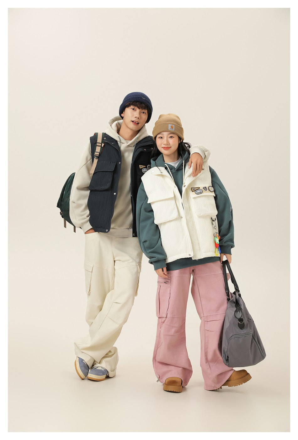 Two models wearing cream and navy utility vests over hoodies, shown full length against a plain background.