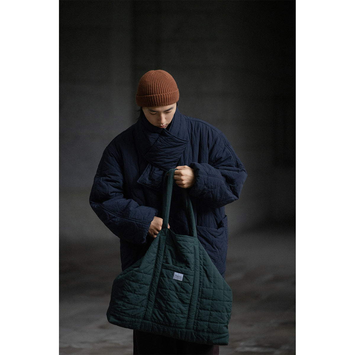 Model wearing a dark navy quilted jacket with a high wrap collar, holding a dark green tote.