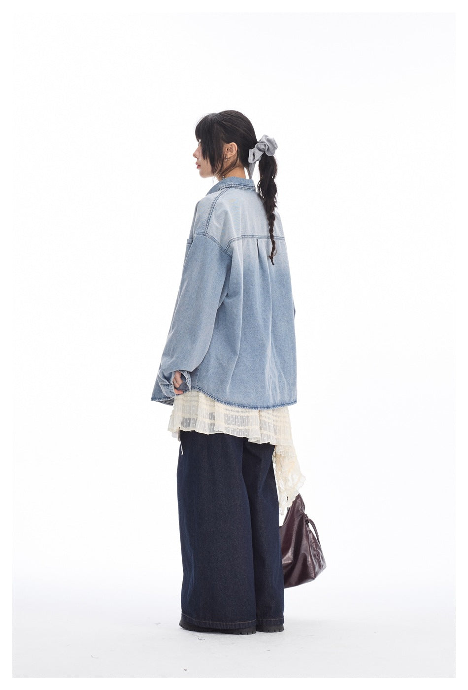 Back view of a model wearing an oversized light blue denim shirt with a yoke seam and center pleat.