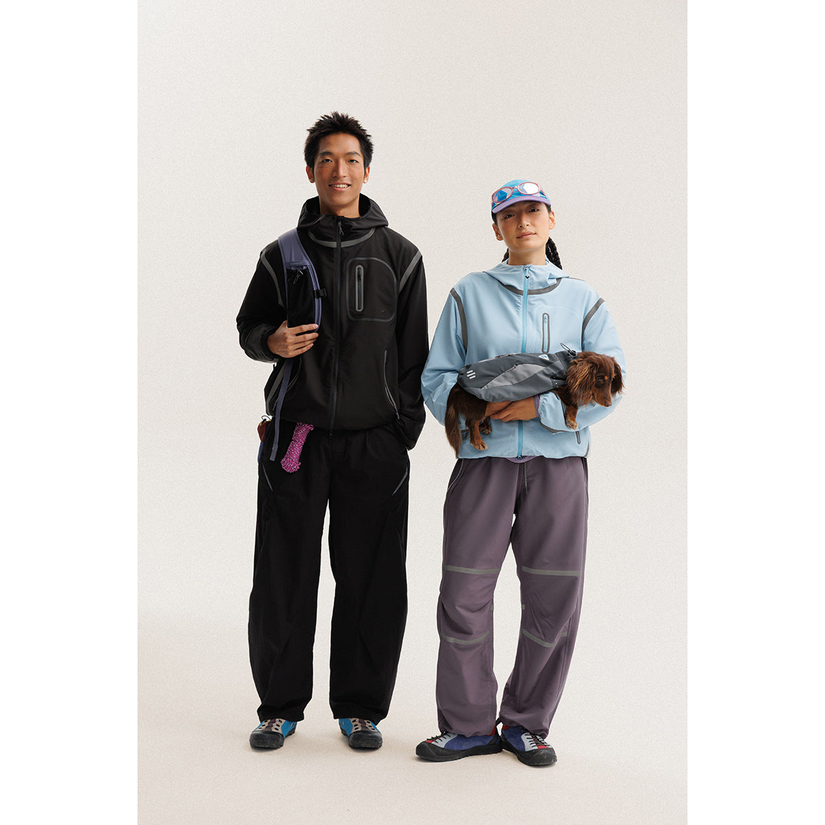 Two models wear black and pale blue hooded jackets with dark outdoor pants; one holds a small dog.