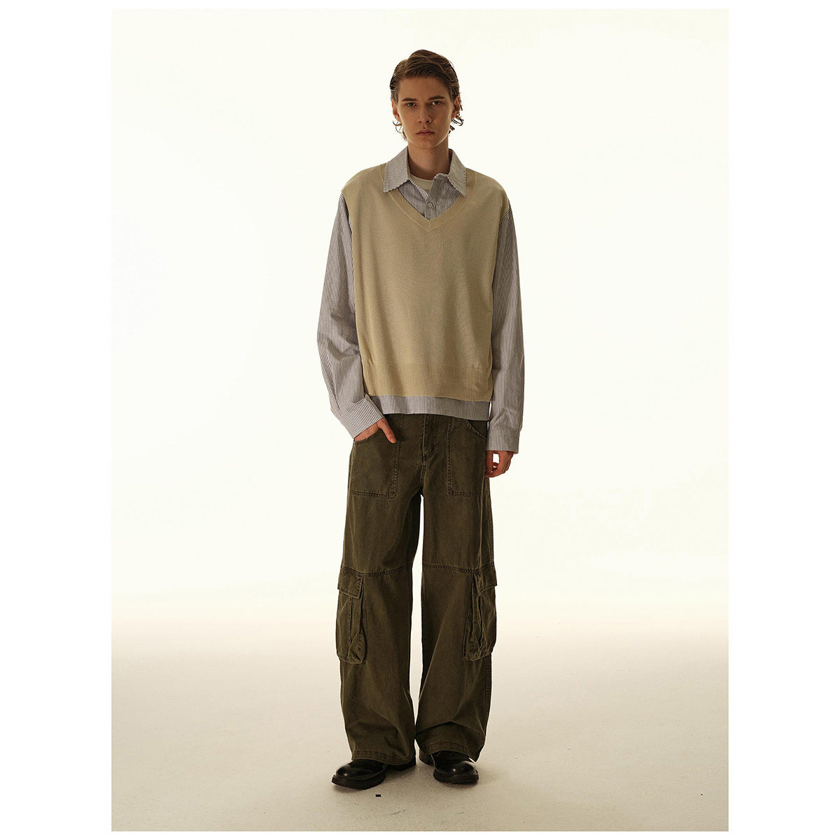 Model wearing a tan V-neck sweater vest over a gray striped shirt and loose dark olive cargo pants.
