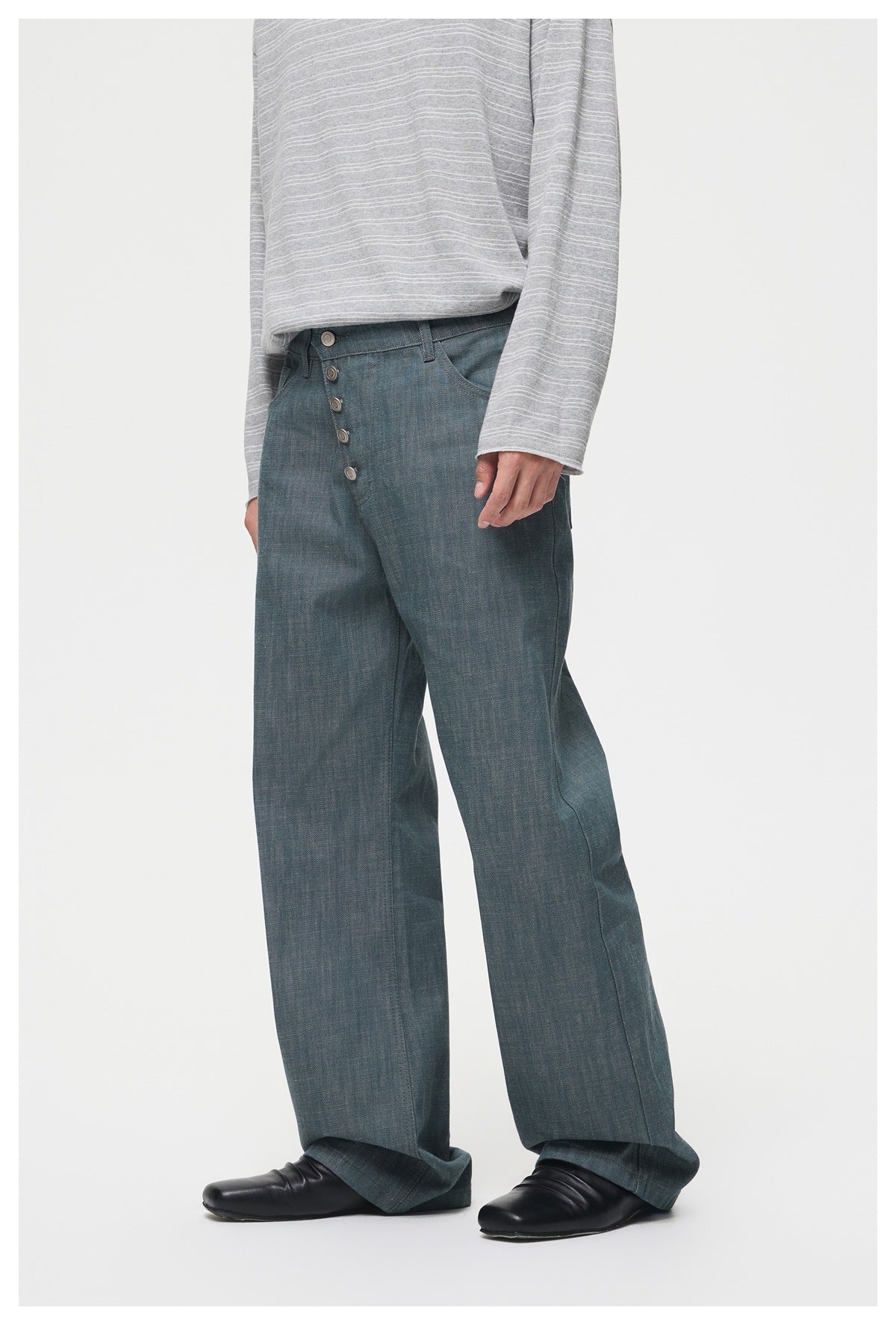 Model wearing loose blue-gray trousers with a button fly, shown from a slight angle with a gray striped top.