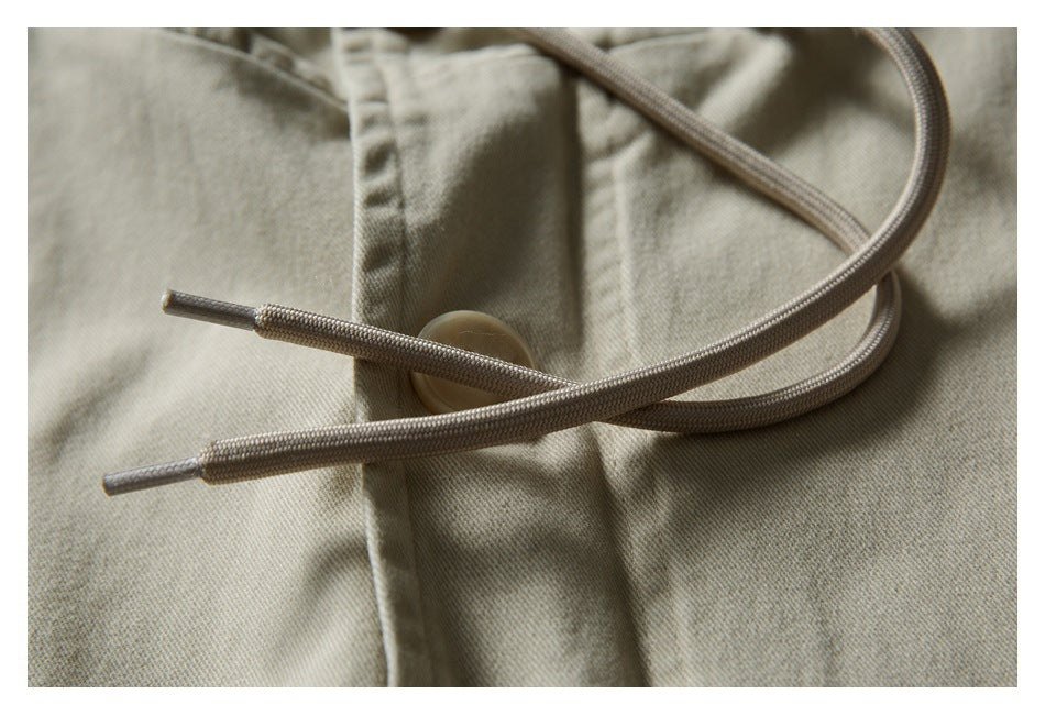 Close-up of braided drawcords crossing a buttoned placket on a light khaki jacket.
