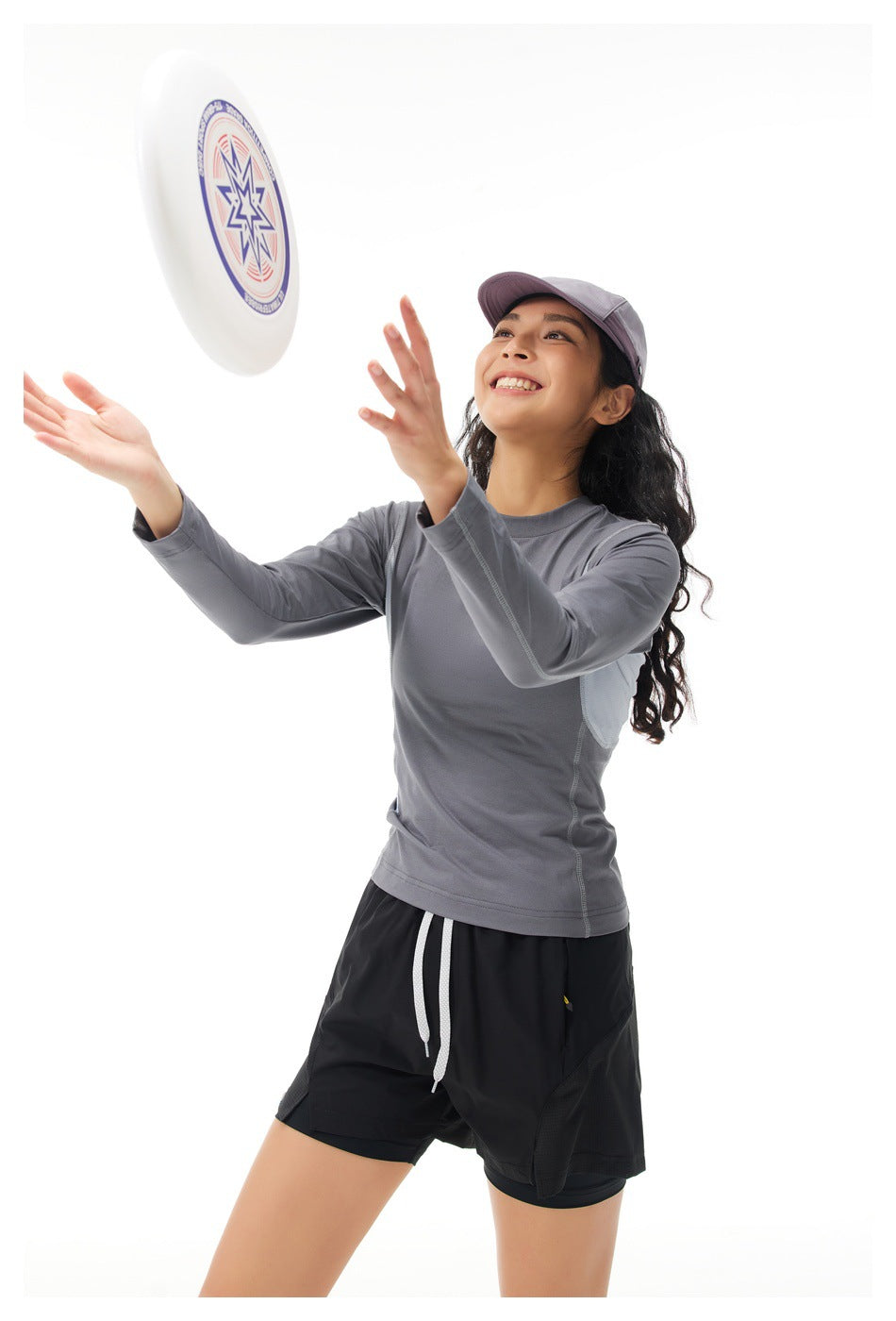 Model in a gray long-sleeve top, black shorts and lavender cap reaching toward a flying disc.
